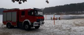 Średni samochód ratowniczo gaśniczy stojący na plaży. W tle ratownicy podczas wykonywania ćwiczeń z deską lodową na plaży jeziora Głębokiego w Szczecinie
