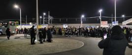 Widok wielu osób stojących z bagażami w nocy na chodniku na terenie dworca kolejowego w Rzepinie. Wśród nich widoczni umundurowani strażacy oraz inne osoby w kamizelkach odblaskowych. Po lewej stronie autobus.