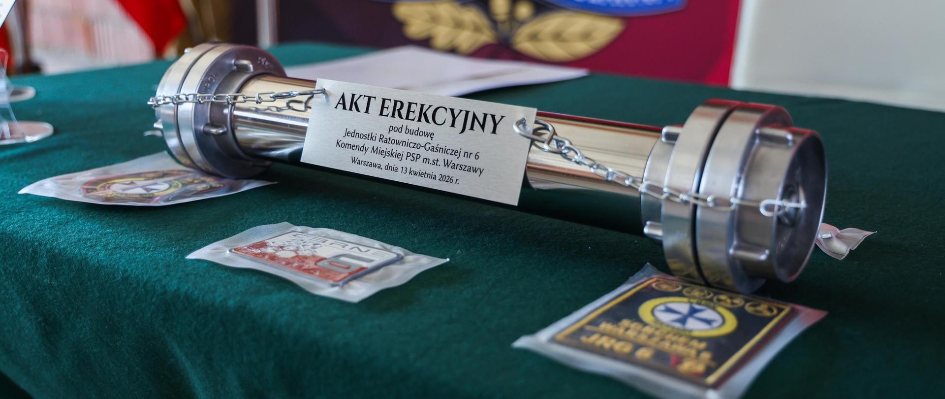 Zbliżenie na metalową tubę z napisem „Akt erekcyjny” oraz pamiątkowe emblematy i monety ułożone na zielonym stole.
