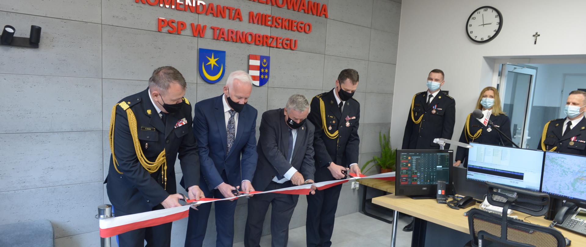 W pomieszczeniu czterech mężczyzn przecina wstęgę biało-czerwoną. Dwóch z mężczyzn to strażacy w umundurowaniu wyjściowym, w tym jeden w umundurowaniu generała, pozostali dwaj to osoby cywilne w garniturach. W pomieszczeniu widoczni są inni strażacy w umundurowaniu. W pomieszczeniu stoi biurko, na którym ustawiono monitory, przy biurku stoi fotel. Na ścianie widoczny napis "STANOWISKO KIEROWANIA KOMENDANTA MIEJSKIEGO PSP W TARNOBRZEGU". 