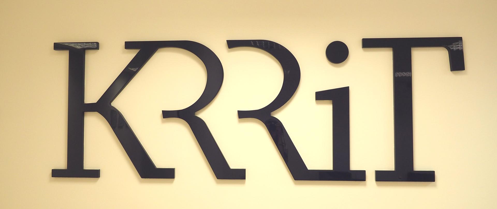Zdjęcie poziome. Czarny napis "KRRiT" na białej ścianie. 