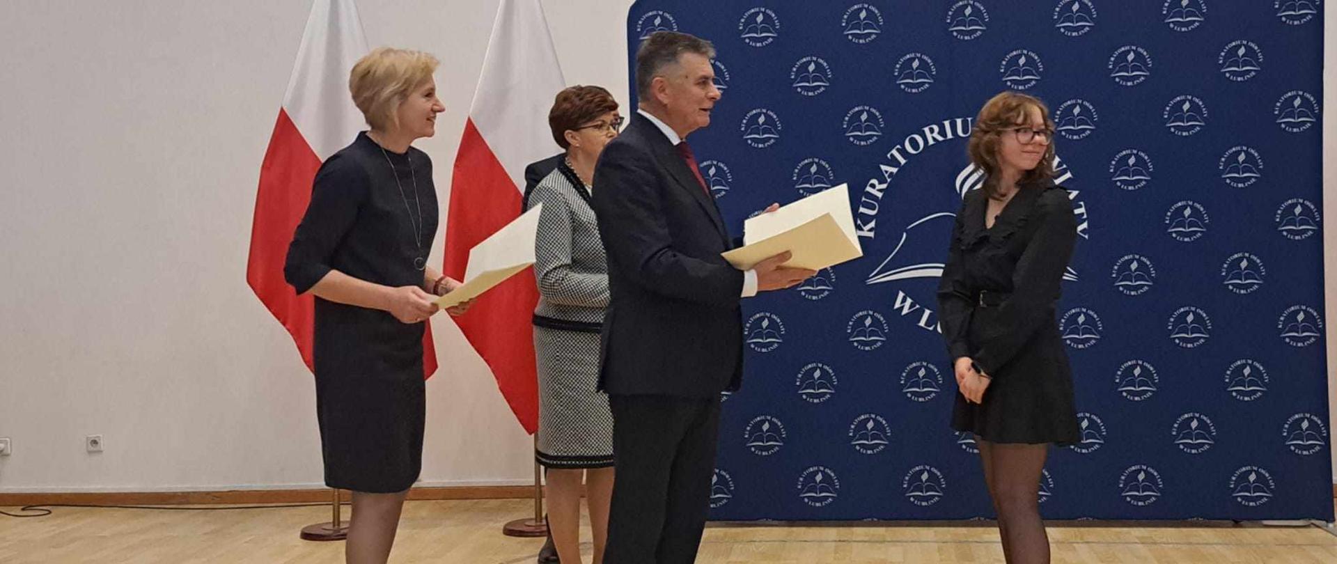 Fotografia. W Sali Błękitnej Urzędu Wojewódzkiego w Lublinie Lech Sprawka składa gratulacje Zuzannie Mereście. Trzyma on w rękach papierowy arkusz zgięty w pół. Za nim stoi młoda kobieta również ma w rękach papierowy arkusz zgięty w pół. Obok nich stoi Teresa Misiuk. W tle na granatowym tle kompzycja: duże logo Kuratorium Oświaty w Lublinie w otoczniu małych logo KO w Lublinie oraz dwie flagi Polski.