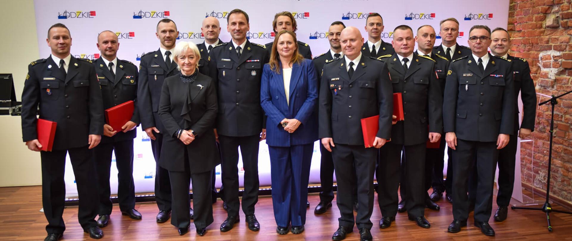 Grupa mężczyzn w mundurach służb mundurowych oraz dwie kobiety w strojach wizytowych, pozująca na tle baneru z napisem "URZĄD MARSZAŁKOWSKI WOJEWÓDZTWA ŁÓDZKIEGO" i logo Łódzkiego. Wszyscy mężczyźni trzymają czerwone teczki.