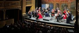 A chamber orchestra on stage conducted by Christoph Koncz. Women are wearing red dresses, men in black suits, white shirts, and pink ties. In the background, roll-ups of the Polish Embassy in Valletta and the De Vallette Chamber Orchestra are visible. The photo is taken from the side and at a distance, showing a few shadowed rows of the audience.