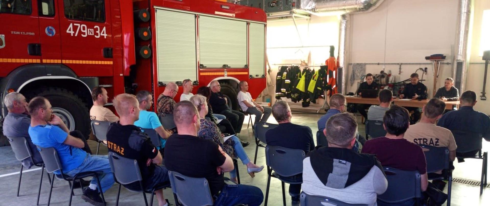 Zdjęcie robione w garażu OSP w Reszlu. Osoby biorące udział w konsultacjach siedzą na krzesłach. Przy stole siedzi dwóch funkcjonariuszy ubranych w czarny Koszar Dowódczo Sztabowy. W tle czerwony wóz bojowy straży pożarnej.