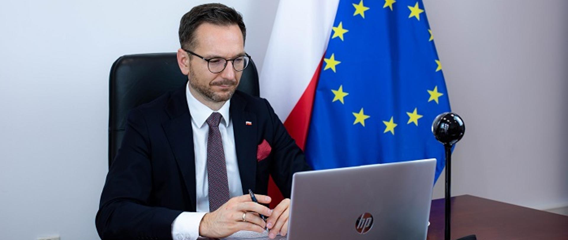 Minister Waldemar Buda siedzi przed laptopem, przy biurku. W dłoni trzyma długopis. Za plecami stoją flagi Polski i UE