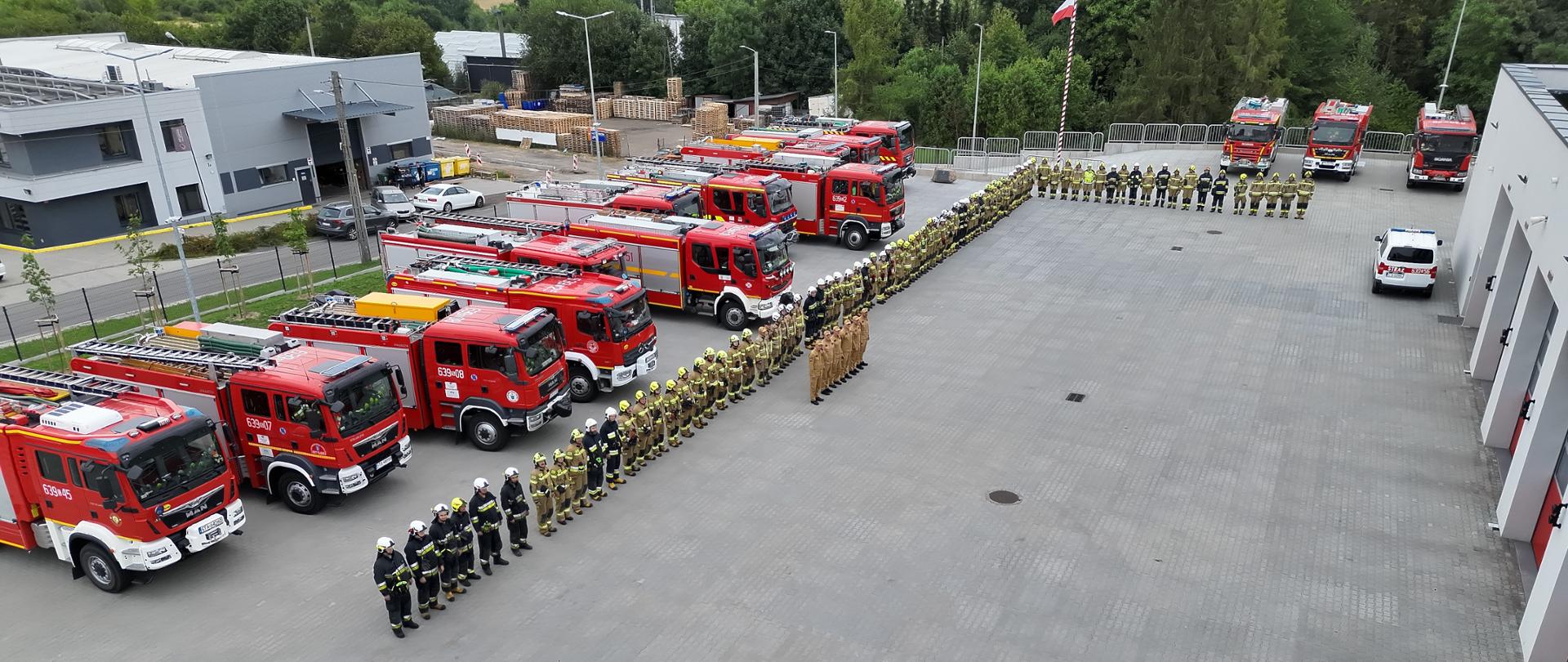 Na zdjęciu widoczny jest plac apelowy przed nowoczesną remizą strażacką.
Kluczowe elementy:
Zbiórka strażaków: Na placu ustawieni są w szyku strażacy – częściowo w ciemnych mundurach galowych/koszarowych, a częściowo w piaskowych/musztardowych mundurach specjalnych (używanych podczas akcji ratowniczo-gaśniczych).
Pojazdy ratowniczo-gaśnicze: Po lewej stronie oraz w tle zaparkowany jest szereg czerwonych wozów strażackich (m.in. marki MAN) oraz pojazdów pomocniczych.
Otoczenie: Po prawej stronie znajduje się budynek jednostki z bramami wyjazdowymi dla wozów, a w tle widać flagę państwową na maszt oraz zielone, leśne tereny.