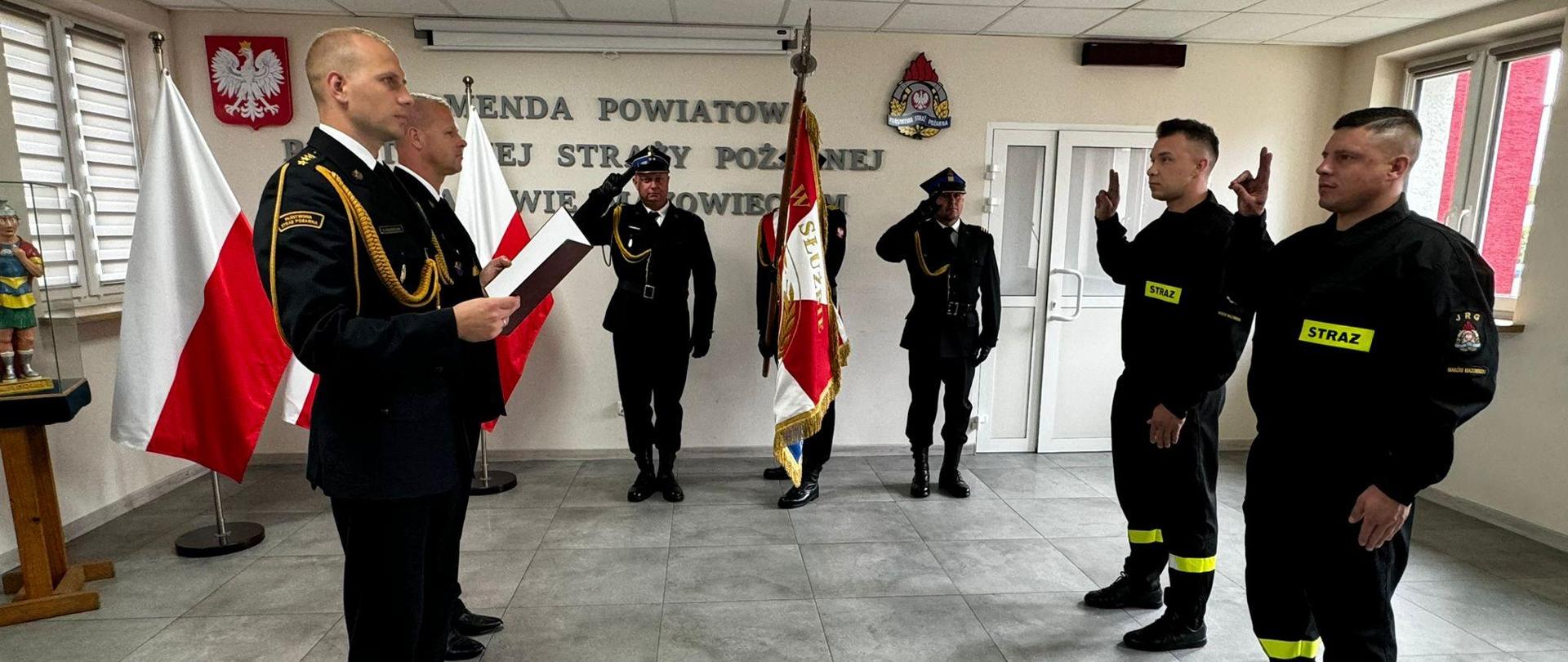 Uroczystość ślubowania funkcjonariuszy na świetlicy Komendy. Dwóch funkcjonariuszy, stojących naprzeciwko Komendantów, podnosi prawą rękę z dwoma palcami uniesiony ku górze. Po środku stoi poczet sztandarowy. 