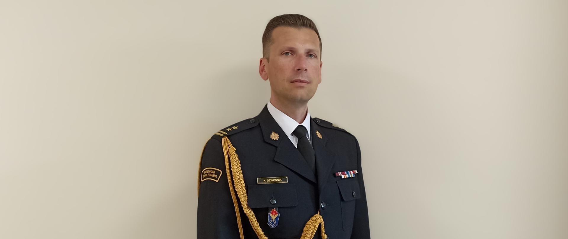Zdjęcie przedstawia bryg. Kamil Dzwonnik Zastępcę Komendanta Miejskiego Państwowej Straży Pożarnej w Częstochowie w mundurze galowym na szarym tle.