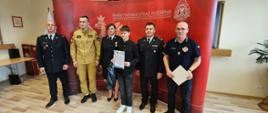 fotografia przedstawia laureata konkursu wraz z kadrą oraz egzaminatorami konkursu, w tle ścianka Państwowa Straż Pożarna, część wyposażenia Sali