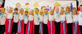 Grupa elegancko ubranych dzieci stoi pod ścianą, na której wisi napis Witamy w nowym przedszkolu.