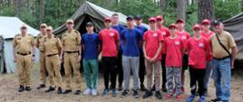 Przed namiotami harcerskimi stoi w szeregu grupa młodzieży oraz mężczyźni ubrani w mundury Państwowej i Ochotniczej Straży Pożarnej - zdjęcie grupowe