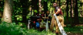 Fotorealistyczne ujęcie z drona na leśną polanę. Na pierwszym planie widać dach czerwonego wozu strażackiego o numerach "89s 699", na którym stoi strażak, z góry rozwijając wąż gaśniczy (odniesienie do pliku D5N_7351.jpg). Na polanie, wokół centralnie stojącego quada (jak w pliku D5N_7392.jpg), zbiórkę odbywa grupa kilkudziesięciu strażaków w piaskowych mundurach służbowych, ustawionych w półkolu (odniesienie do formacji z pliku D5N_7351.jpg). Dwóch strażaków w czerwonych mundurach bojowych w środku grupy wskazuje na quada i węże, koordynując działania. W tle gęsty, dojrzały las i zaparkowane inne pojazdy strażackie. Scena dokumentuje dynamiczne ćwiczenia z rozwijania linii gaśniczych w terenie leśnym. W prawym dolnym rogu widoczny jest logotyp PSP.
