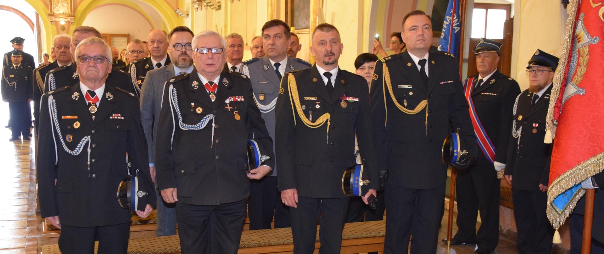 na głównym planie czterech strażaków w mundurach wyjściowych 2 ochotników i 2 strażaków zawodowych, stoją w kościele przy pierwszej ławce, za nimi inne osoby i strażacy
