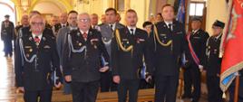 na głównym planie czterech strażaków w mundurach wyjściowych 2 ochotników i 2 strażaków zawodowych, stoją w kościele przy pierwszej ławce, za nimi inne osoby i strażacy
