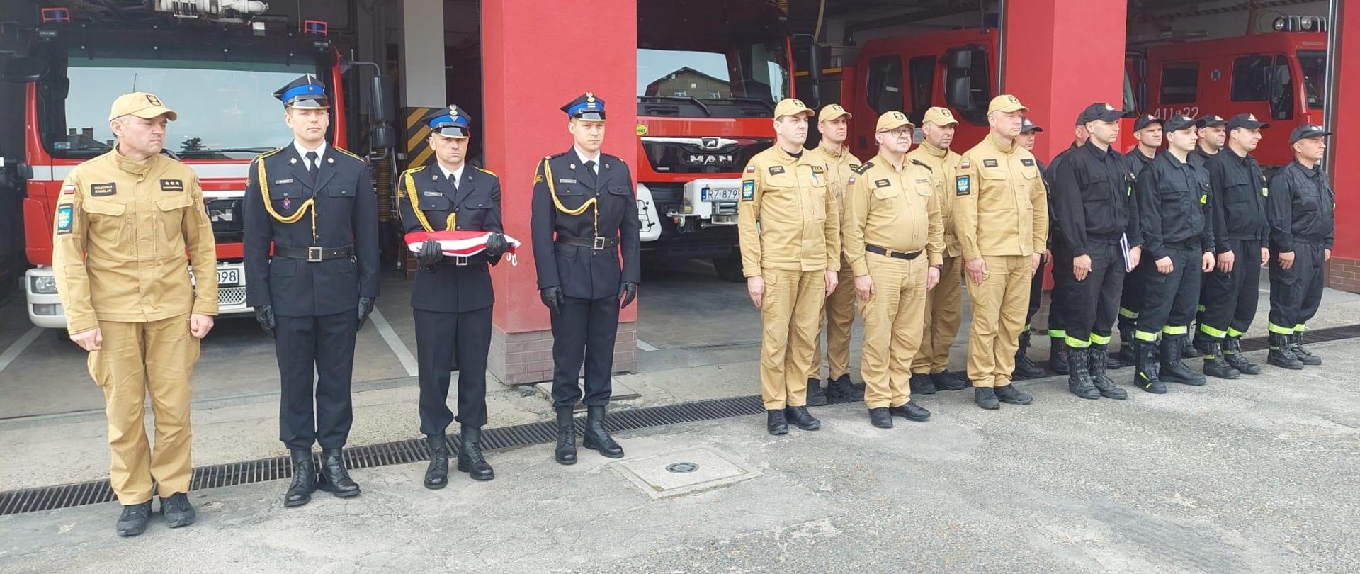 Kolorowa fotografia wykonana na zewnątrz w pochmurny dzień. Przedstawia strażaków w mundurach służbowych i koszarowych przed strażnicą PSP w Brzozowie. Na pierwszym planie przygotowany poczet flagowy w mundurach wyjściowych z flagą na ręku.