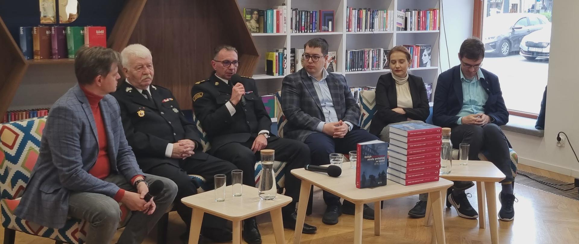 Sześć osób siedzi na panelu dyskusyjnym w nowoczesnej bibliotece. Dwóch mężczyzn w mundurach strażackich trzyma mikrofony, a pozostali uczestnicy są ubrani formalnie. Na stolikach przed nimi stoją szklanki, karafki z wodą oraz stos czerwonych książek. W tle widoczne są regały z książkami i zielona tabliczka z napisem „Nowości”, a po prawej stronie duże okno wychodzące na ulicę.