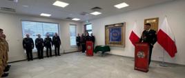 Widzimy uroczystą, oficjalną zbiórkę w budynku straży pożarnej. Na środku stoi mężczyzna w galowym mundurze, przemawiający z mównicy ozdobionej godłem Państwowej Straży Pożarnej. Po jego bokach ustawiono dwie polskie flagi. Wzdłuż lewej ściany stoi grupa strażaków w mundurach — część w ciemnych galowych, część w jasnych, polowych. Z tyłu sali znajduje się kilku gości lub przełożonych obserwujących wydarzenie. Pomieszczenie jest jasne, nowoczesne, z białymi ścianami i płytkami na podłodze, co podkreśla formalny charakter ceremonii.