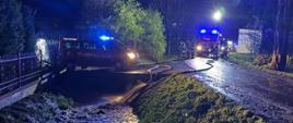 Nocna akcja strażaków PSP przy wezbranym potoku; pojazdy pożarnicze z włączonymi światłami i rozwinięte węże na zalanej drodze.