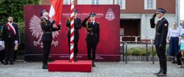 Fotografia przedstawia uroczysty moment podnoszenia flagi państwowej na maszt przez strażaków Państwowej Straży Pożarnej. Ceremonia odbywa się na placu przed budynkiem, na tle czerwonej ścianki promocyjnej z godłem Polski i logo formacji. Strażacy w galowych mundurach salutują oraz wciągają biało-czerwoną flagę.