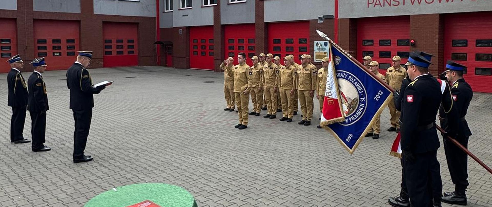 Na zdjęciu przedstawiono uroczystą zbiórkę funkcjonariuszy Państwowej Straży Pożarnej na placu Komendy Miejskiej PSP. W ceremonii uczestniczą strażacy ustawieni w szyku, poczet sztandarowy oraz kadra kierownicza prowadząca uroczystość.
W centralnej części placu znajduje się stół z dokumentami przygotowanymi do podpisania lub wręczenia. W tle widoczny jest budynek komendy z garażami dla pojazdów ratowniczo-gaśniczych. Fotografia przedstawia oficjalny przebieg uroczystości związanej z realizacją zadań służbowych Państwowej Straży Pożarnej.
