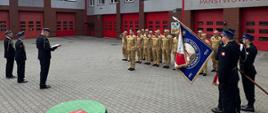 Na zdjęciu przedstawiono uroczystą zbiórkę funkcjonariuszy Państwowej Straży Pożarnej na placu Komendy Miejskiej PSP. W ceremonii uczestniczą strażacy ustawieni w szyku, poczet sztandarowy oraz kadra kierownicza prowadząca uroczystość.
W centralnej części placu znajduje się stół z dokumentami przygotowanymi do podpisania lub wręczenia. W tle widoczny jest budynek komendy z garażami dla pojazdów ratowniczo-gaśniczych. Fotografia przedstawia oficjalny przebieg uroczystości związanej z realizacją zadań służbowych Państwowej Straży Pożarnej.