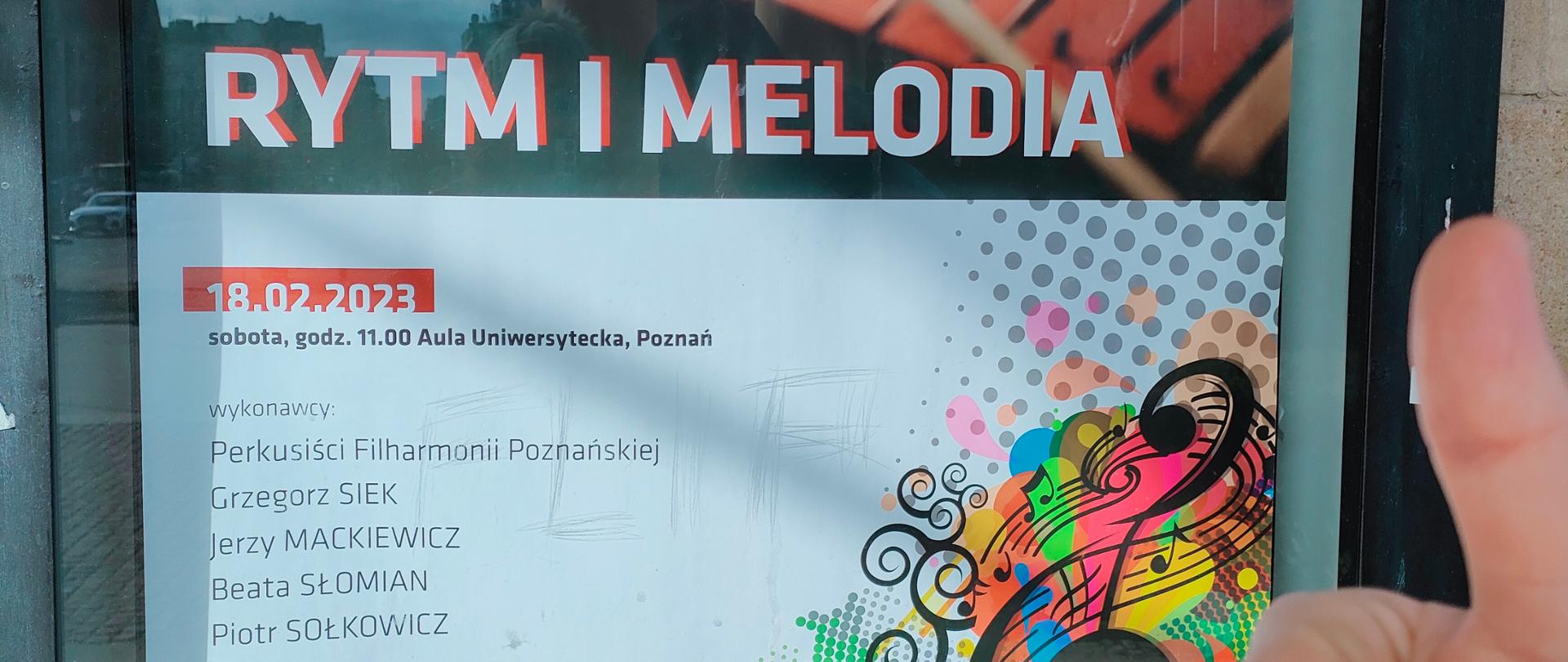 Zdjęcie prezentuje zdjęcie plakatu informacyjnego o koncercie perkusyjnym w witrynie Filharmonii Poznańskiej. Z prawej strony widnieje uniesiony w górę kciuk dłoni ucznia. 