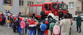 Na zdjęciu widać grupę przedszkolaków, biorących udział w pokazie prowadzonym przez strażaków. Za dziećmi stoi wóz strażacki i trzech strażaków. Pokaz odbywa się na zewnątrz na placu przy budynku przedszkola