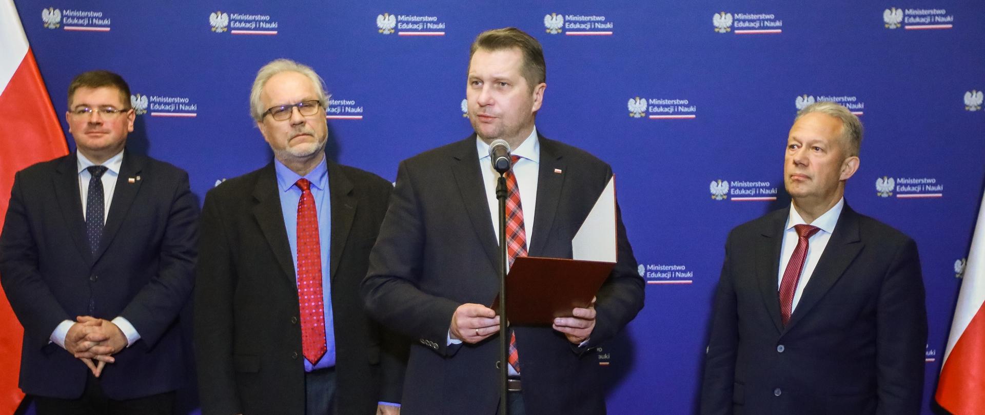 Czterech mężczyzn w garniturach stoi przed niebieską ścianą, za nimi po bokach polskie flagi, wśród nich minister Czarnek który mówi do stojącego na stojaku mikrofonu i trzyma w ręku czerwoną teczkę.