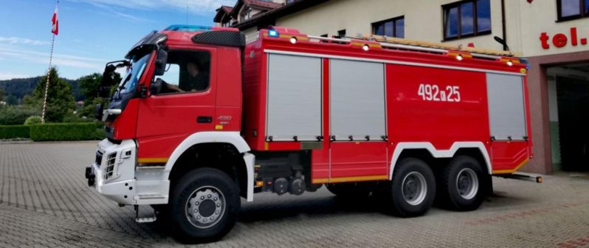 Na zdjęciu jest czerwony samochód strażacki na tle remizy w Rabce-Zdroju. na burcie auta znajduje się numer operacyjny 492-25. na drugim planie jest maszt z flagą państwową, z niebo jest zasnute chmurami strzępiastymi.