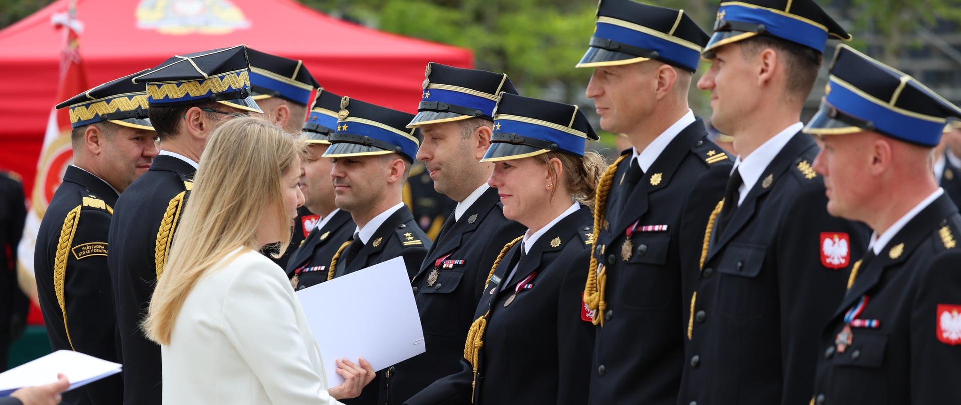 Na pierwszym planie kobieta w jasnym, kremowym żakiecie wręcza dokument i ściska dłoń funkcjonariuszce stojącej w szeregu. Po obu stronach ustawieni są umundurowani funkcjonariusze w granatowych mundurach galowych z odznaczeniami, złotymi sznurami i czapkami z niebieskimi otokami. Kilka osób trzyma białe teczki lub dyplomy. Na mundurach widoczne są czerwono-białe emblematy z wizerunkiem białego orła. W tle znajduje się czerwony namiot i zieleń drzew. Zdjęcie przedstawia moment oficjalnego wręczania wyróżnień, nominacji lub odznaczeń podczas uroczystości służbowej.