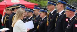 Na pierwszym planie kobieta w jasnym, kremowym żakiecie wręcza dokument i ściska dłoń funkcjonariuszce stojącej w szeregu. Po obu stronach ustawieni są umundurowani funkcjonariusze w granatowych mundurach galowych z odznaczeniami, złotymi sznurami i czapkami z niebieskimi otokami. Kilka osób trzyma białe teczki lub dyplomy. Na mundurach widoczne są czerwono-białe emblematy z wizerunkiem białego orła. W tle znajduje się czerwony namiot i zieleń drzew. Zdjęcie przedstawia moment oficjalnego wręczania wyróżnień, nominacji lub odznaczeń podczas uroczystości służbowej.