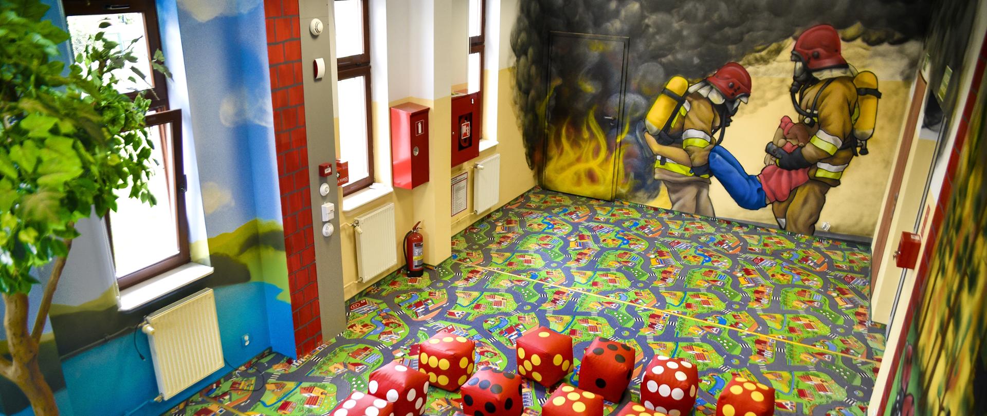 Sala edukacyjna dla dzieci. Na dywanie pufy w kształcie kostki do grania, na ścianach namalowanych jest dwóch strażaków wynoszących osobę poszkodowaną. Na podłodze znajduje się kolorowa wykładzina. 