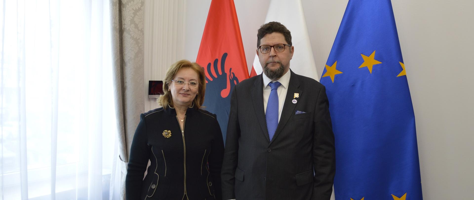 Wiceminister Szeptycki stoi obok kobiety w czarnym ubraniu, za nimi pod białą ścianą flagi Polski, Albanii i UE, w lewej zasłonięte firankami okno.