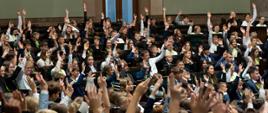 Grupa uczniów i uczennic siedzi w ławach sejmowych i bierze udział w głosowaniu poprzez podniesienie ręki do góry.