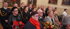 W pierwszym planie siedzą kobiety i mężczyźni, część z nich trzyma bukiety kwiatów w czerwonych i zielonych barwach. W tle stoją i siedzą funkcjonariusze PSP w mundurach galowych. Twarze uczestników są skupione i spokojne. Sala jest jasna, oświetlona światłem dziennym, a na ścianach widoczne są portrety dawnych postaci.