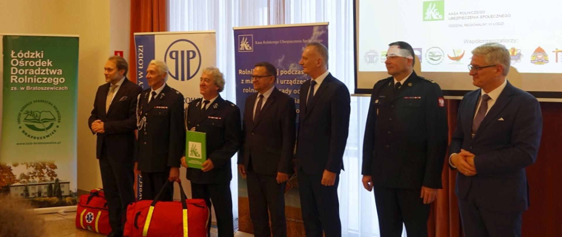 siedmiu mężczyzn stojących w szeregu wśród nich dwóch posłów na Sejm RP dwóch druhów osp oraz zastępca komendanta wojewódzkiego psp, druhowie i strażak są w mundurach wyjściowych, pozostali mężczyźni w garniturach, w tle rollupy różnych instytucji