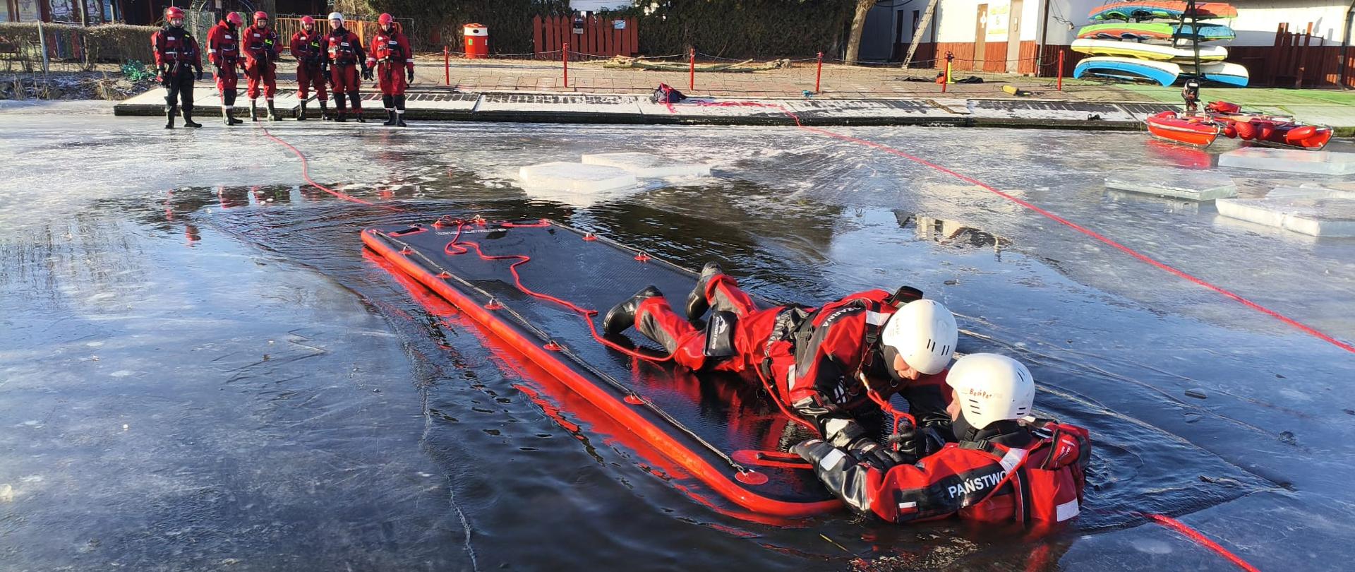 Szkolenie doskonalące w zakresie ratownictwa lodowego. Strażacy ubrani w skafandry nurkowe podczas szkolenia na zamarzniętym akwenie wodnym, ćwiczą wykorzystanie sprzętu do ratownictwa lodowego oraz technik ratownictwa lodowego i samoratownictwa.