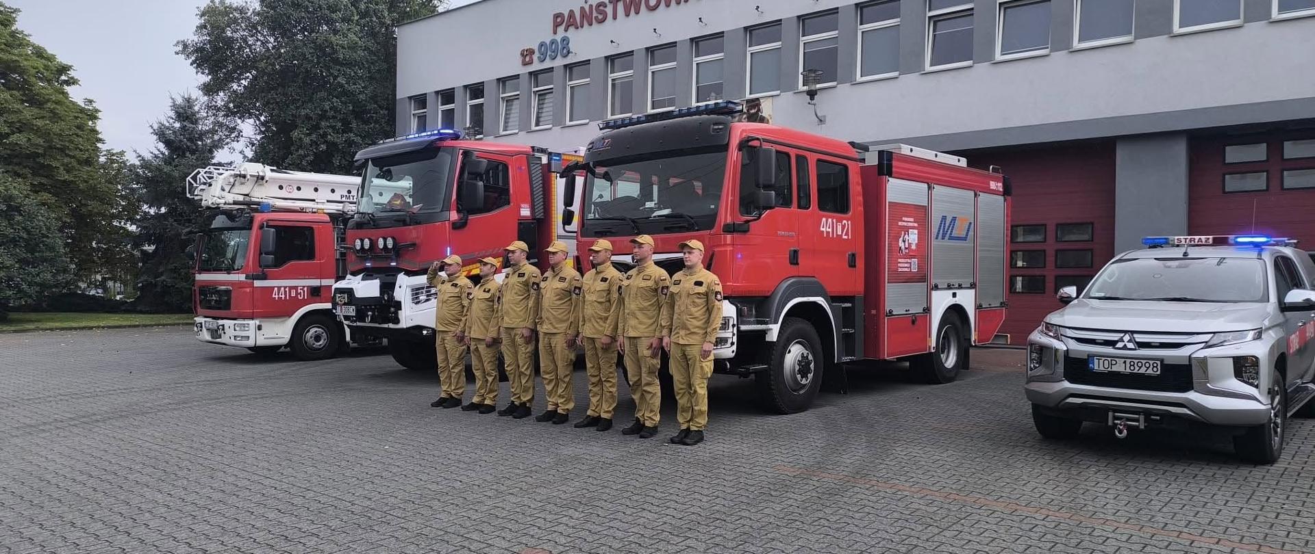 Strażacy Państwowej Straży Pożarnej stoją w szeregu przed budynkiem Komendy Powiatowej PSP. Na placu ustawione są pojazdy pożarnicze, w tym drabina mechaniczna, samochody ratowniczo-gaśnicze oraz oznakowany samochód operacyjny z włączonymi sygnałami świetlnymi.