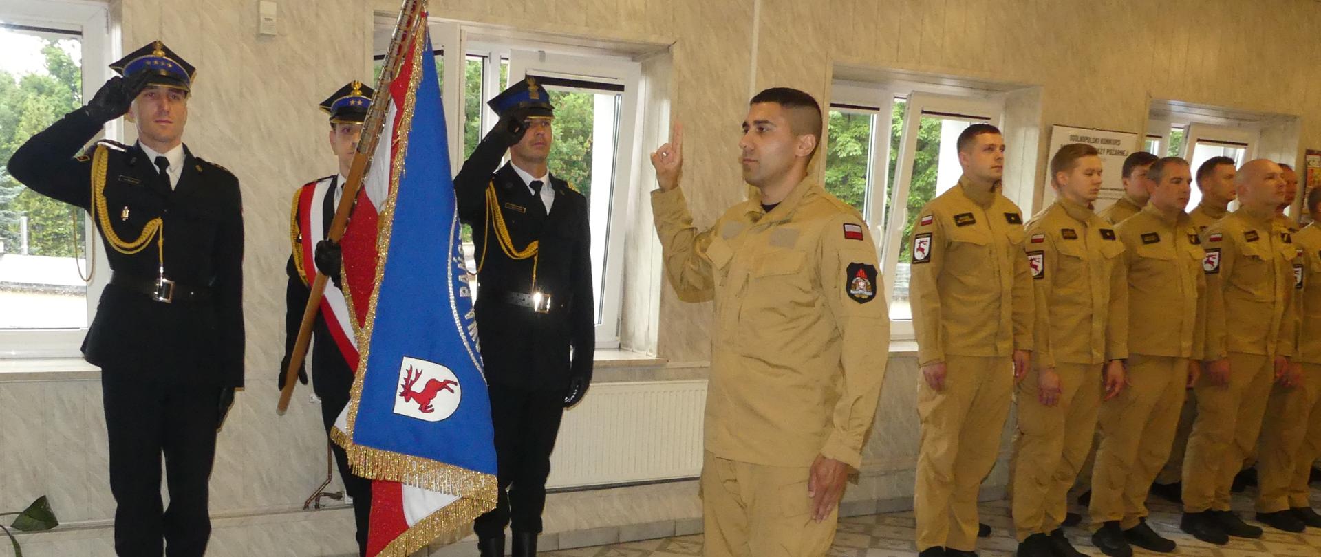 Zdjęcie przedstawia strażaka Adama Asiryana w momencie uroczystego ślubowania. Po lewo od niego stoi poczet sztandarowy Komendy. Młody strażak uniósł prawą dłoń i wypowiada rotę ślubowania w kierunku pochylonego sztandaru. Za nim stoją inni strażacy.