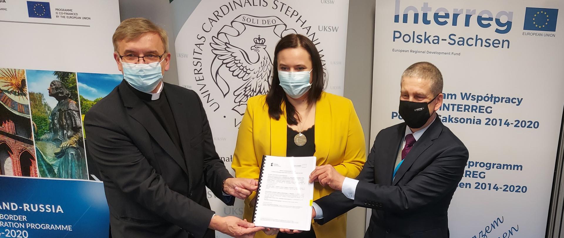 uczestnicy spotkania trzymają przed sobą podpisaną umowę na projekt Sygnalizator Plus dofinansowanego z Funduszy Europejskich