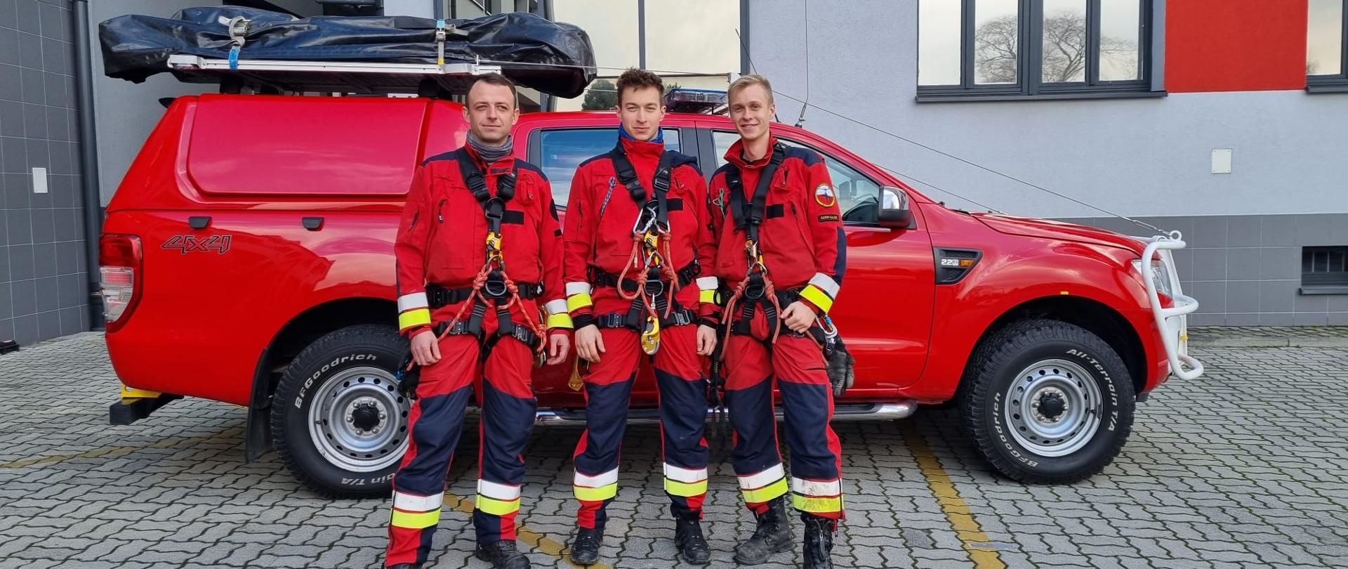 Na zdjęciu widzimy trzech ratowników pozujących do zdjęcia. Ratownicy to mężczyźni w młodym wieku, ubrani w czerwone kombinezony oraz czarne szelki asekuracyjne. Za ratownika znajduje się specjalistyczny samochód ratownictwa wysokościowego koloru czerwonego, ustawiony bokiem do kadru, srebrne felgi. Na dachu samochodu widzimy czarny pokrowiec. Dalej widzimy część budynku koloru szarego z elementami czerwieni. 