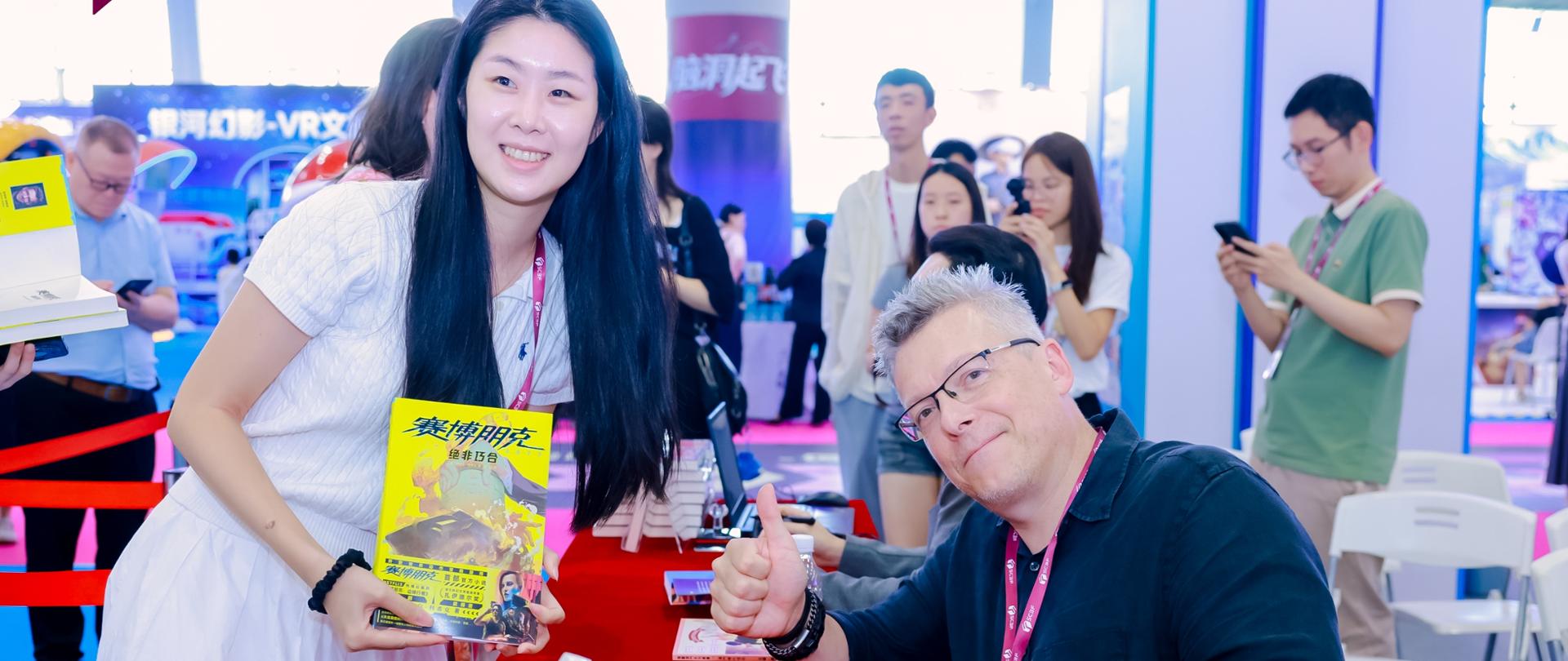 Fanka literatury fantastycznej trzymająca książkę ''Cyberpunk 2077: Bez przypadku'' pozuje do zdjęcia z autorem powieści, Rafałem Kosikiem. Sesja podpisywania autografów przez polskiego autora