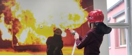 Na zdjęciu dziecko w hełmie czerwonym i z prądownicą strażacką. Przed dzieckiem obraz pożaru, ognia.