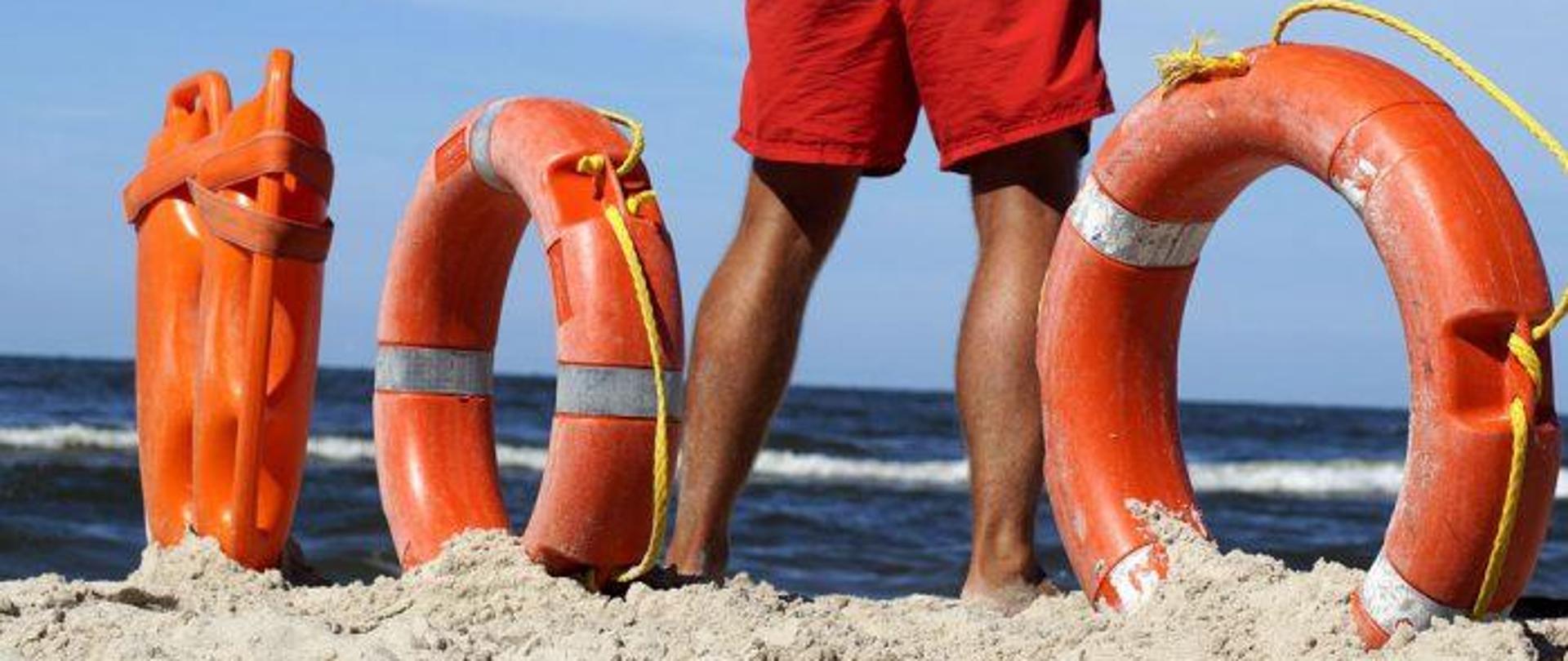 Na rysunku widać plażę i ratownika. Obok niego po jednej stronie i drugiej koła ratunkowe i dwie boje do pływania