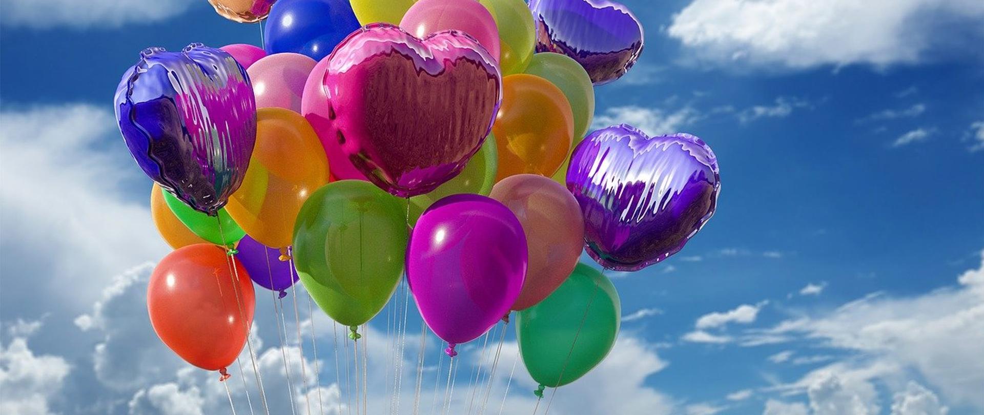 Na tle niebieskiego nieba upstrzonego białymi chmurkami unosi się balonowy bukiet, na który składają się różnokolorowe, okrągłe balony i foliowe balony w kształcie serca. 