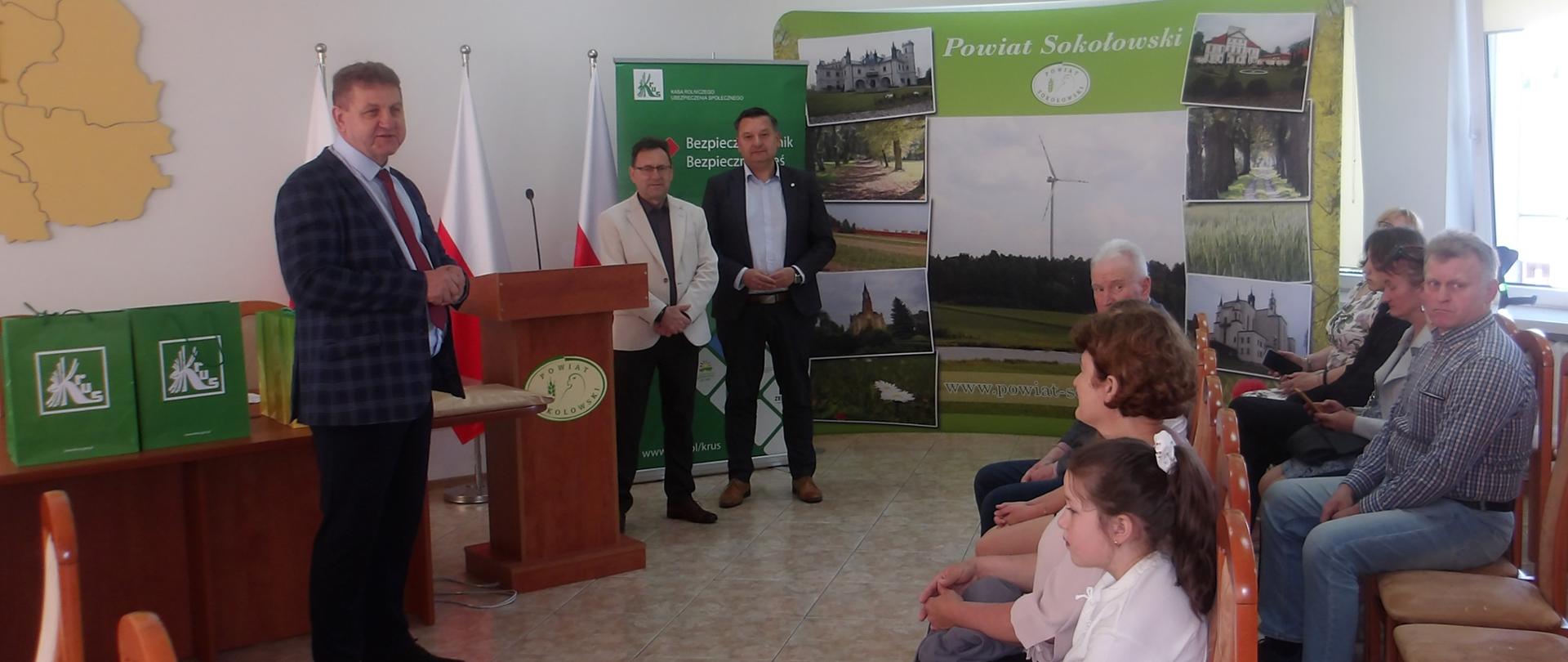 Na sali trzech mężczyzn stoi na tle flag Polski, baneru KRUS i Powiatu Sokołowskiego, po prawej siedzą dzieci z rodzicami.