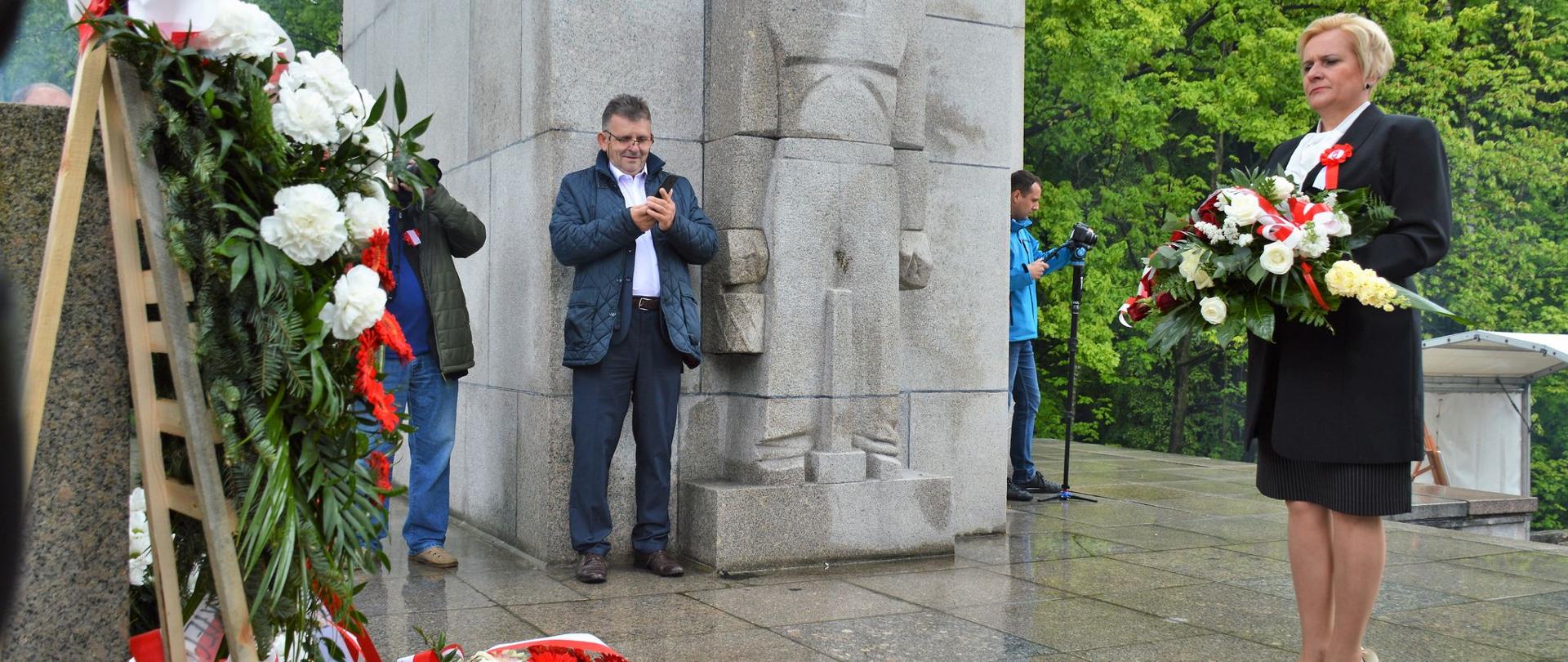 Uroczystości na Górze św. Anny. 22 maja 2021 r. Na zdjęciu wicewojewoda opolski stojąca na przeciw Pomnika na Górze św.Anny. Po pomnikiem floret z biało-czerwonymi kwiatami. W tle dziennikarze,robiący zdjęcia. 