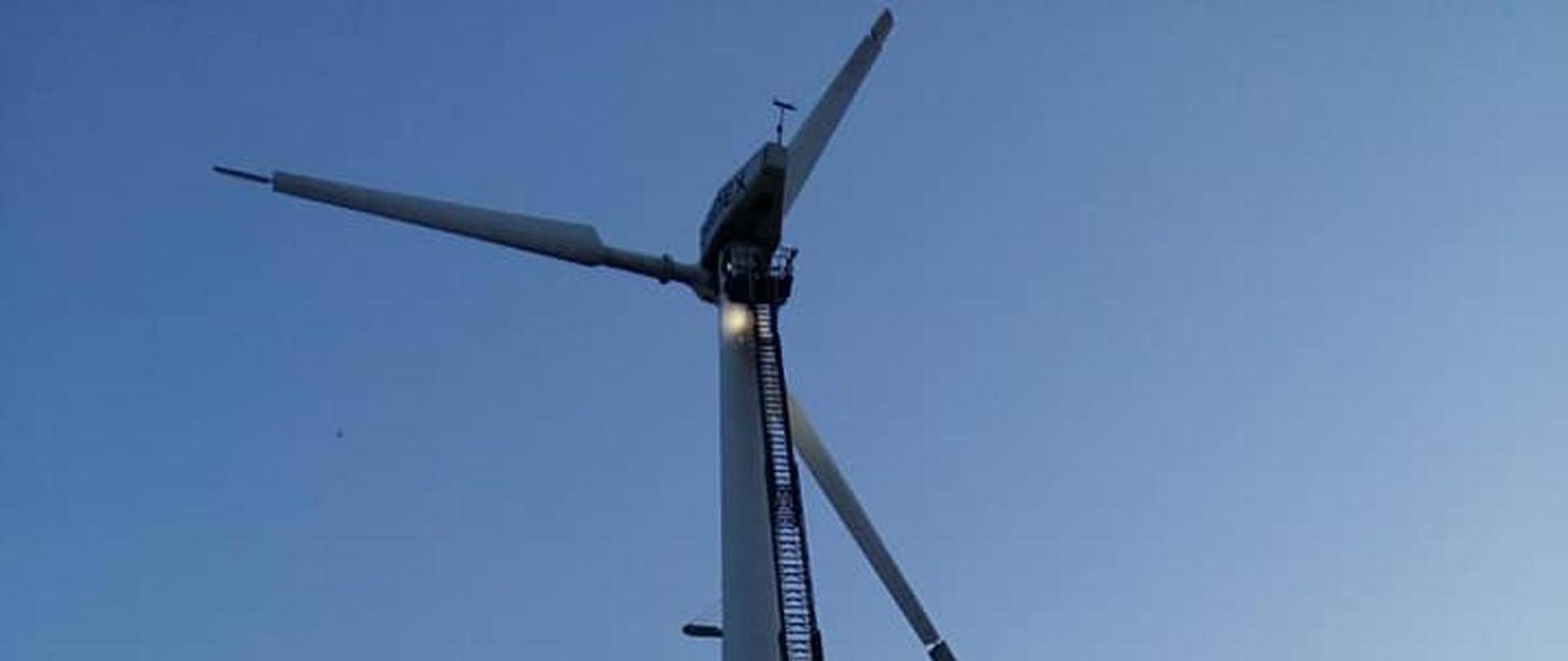 W tle elektrownia wiatrowa. Przy wiatraku rozłożona drabina mechaniczna. Działanie gaśnicze prowadzone przez strażaków podających proszek gaśniczy do wnętrza gondoli z podestu drabiny. Na pierwszym planie dwa samochody pożarnicze.