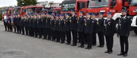 Grupa strażaków w strojach galowych ustawiona w dwuszeregu w trakcie uroczystej zbiórki. W tle samochody pożarnicze.